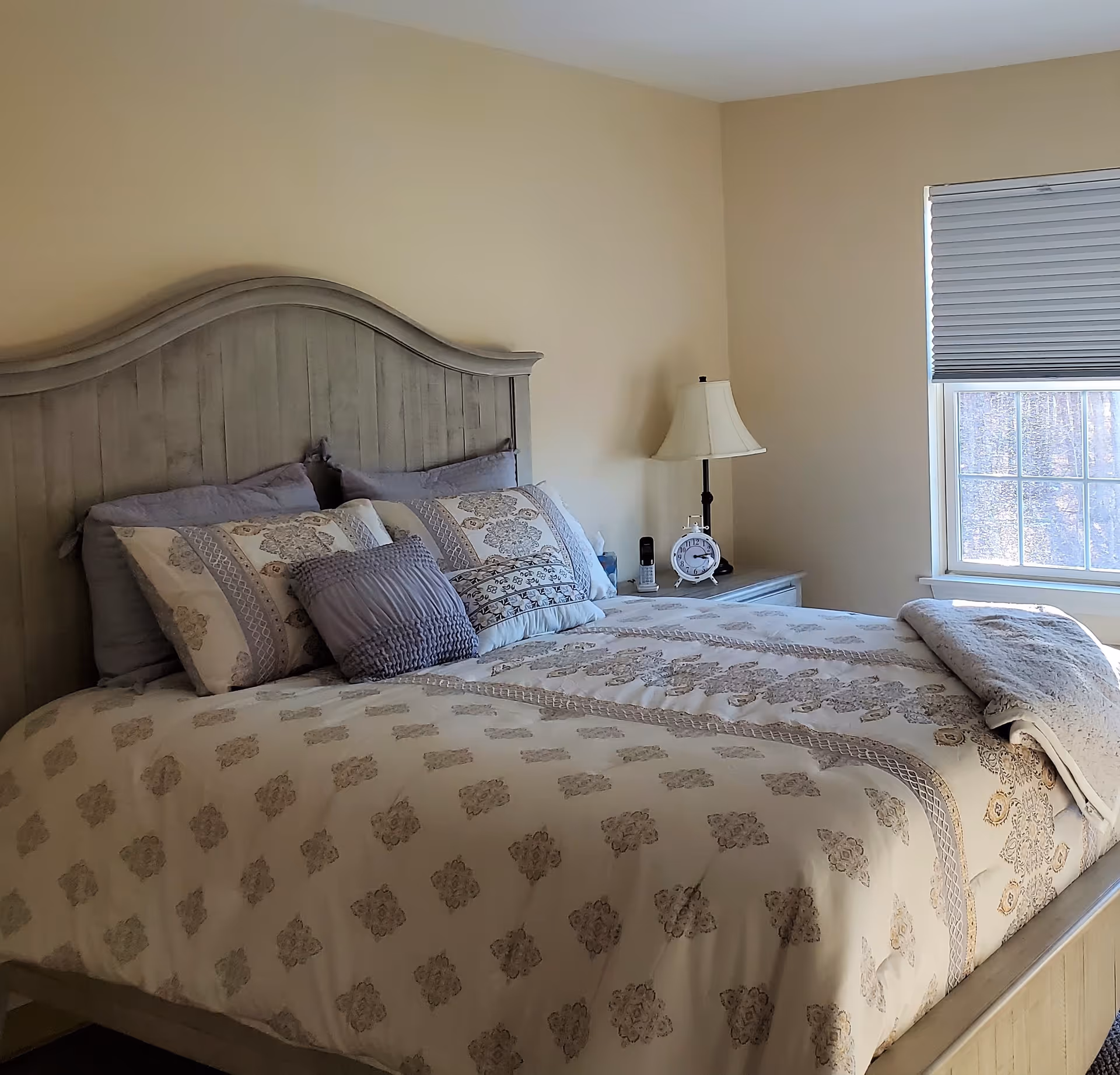 A bedroom with a large wooden bed featuring a patterned beige and gray bedspread and multiple pillows. Next to the bed is a nightstand with a white table lamp, an alarm clock, and a cordless phone. A window with a closed blind allows natural light to enter the room.