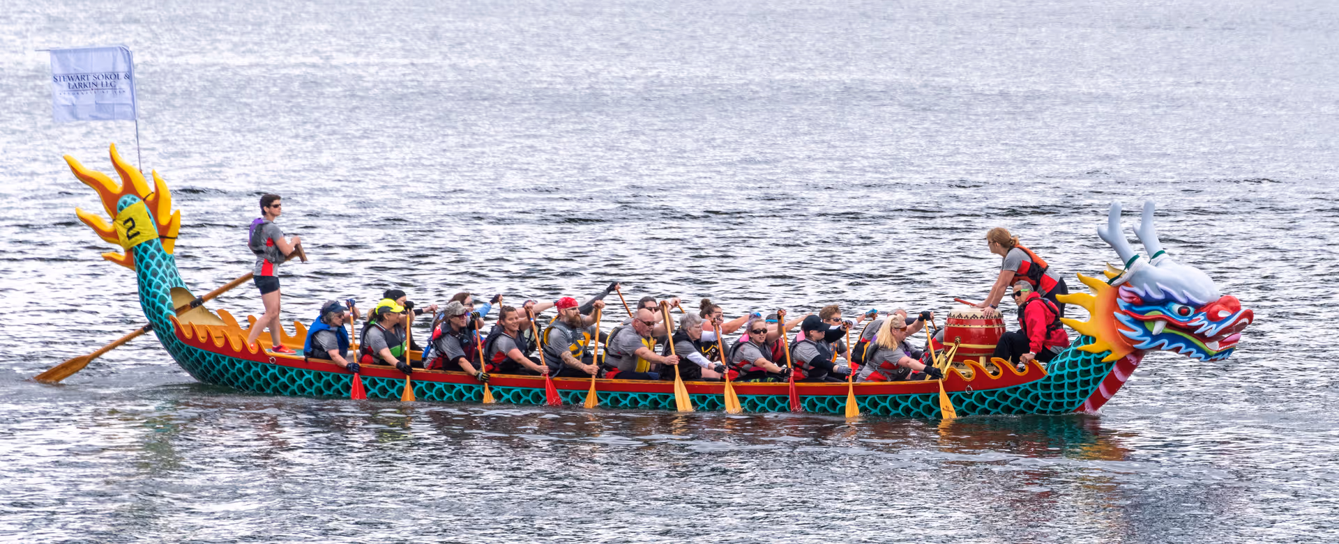 A group of people paddling a colorful dragon boat on a body of water. The boat has a dragon head at the front and a dragon tail at the back. The paddlers are wearing life jackets and are actively rowing. One person stands at the back steering the boat, and another person is positioned at the front near a drum.