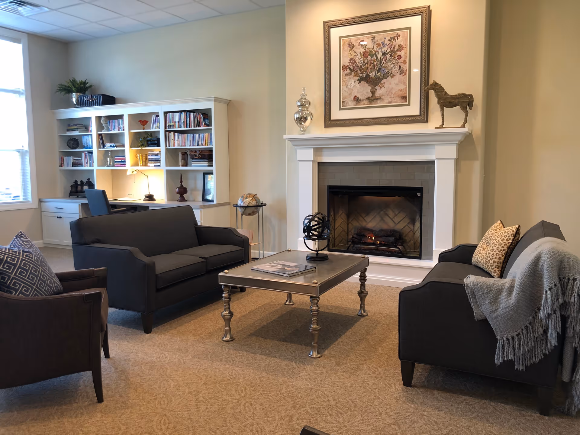 A cozy living room with two dark gray sofas and a brown armchair arranged around a metallic coffee table. There is a fireplace with a white mantel, decorated with a framed floral painting, a silver vase, and a wooden horse sculpture. A built-in white bookshelf and desk with books and decorative items are visible in the background near a window.