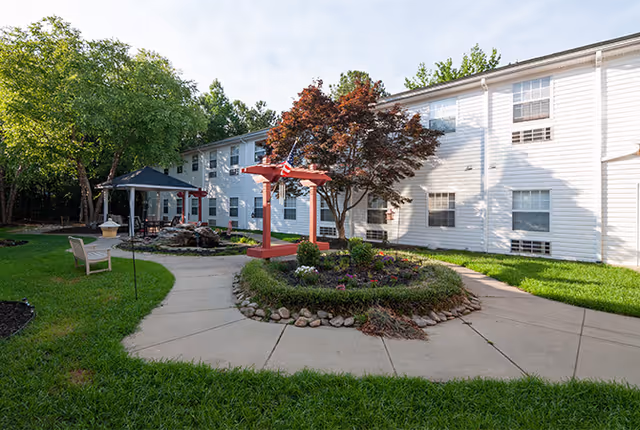 Outdoor courtyard area of a senior living facility with a circular flower bed surrounded by a paved walkway, a red pergola with an American flag, benches, a gazebo, and a two-story white building in the background.