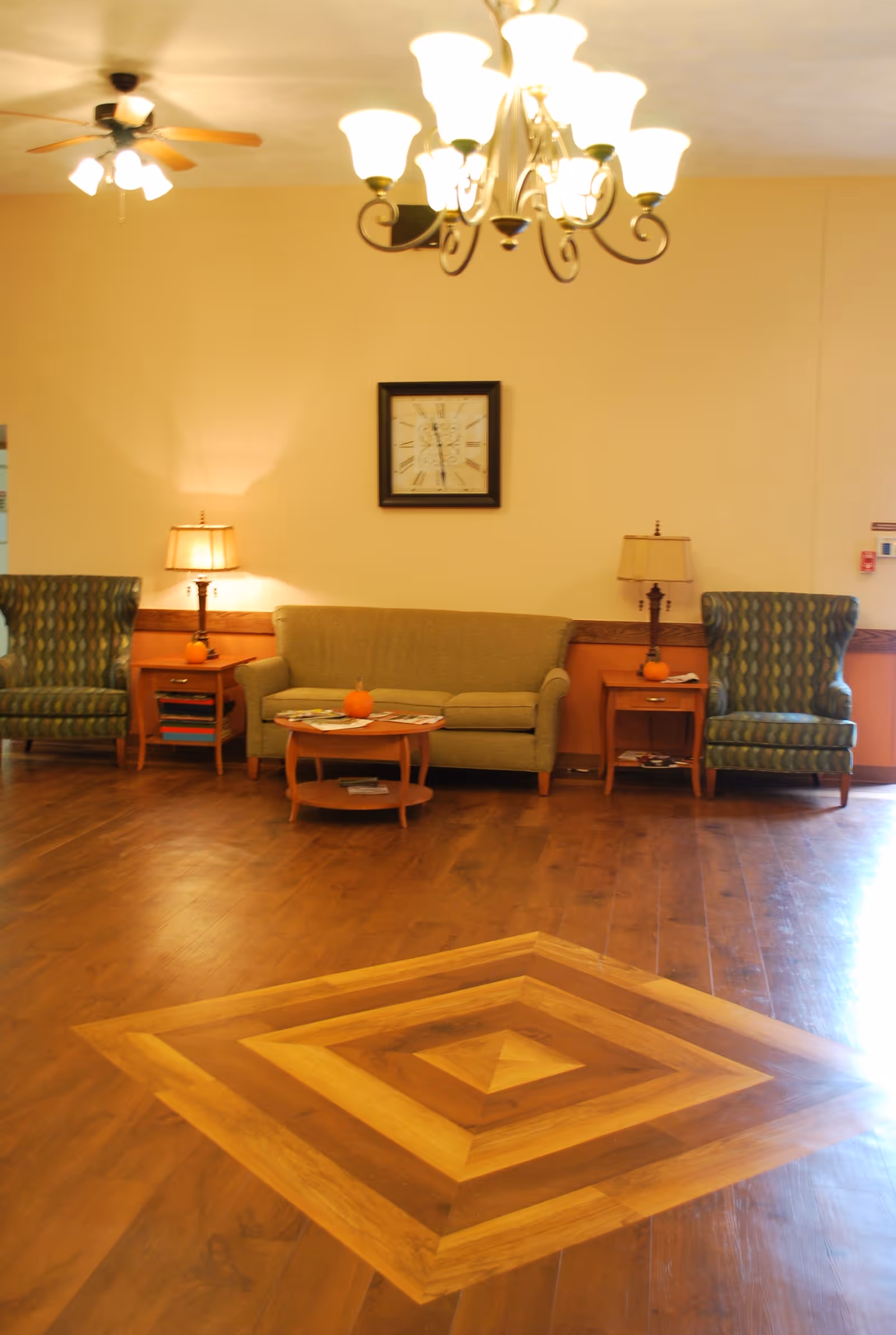 A cozy sitting area with a sofa and two armchairs against a wall, lamps and a clock above, and a decorative wooden floor medallion in the foreground.