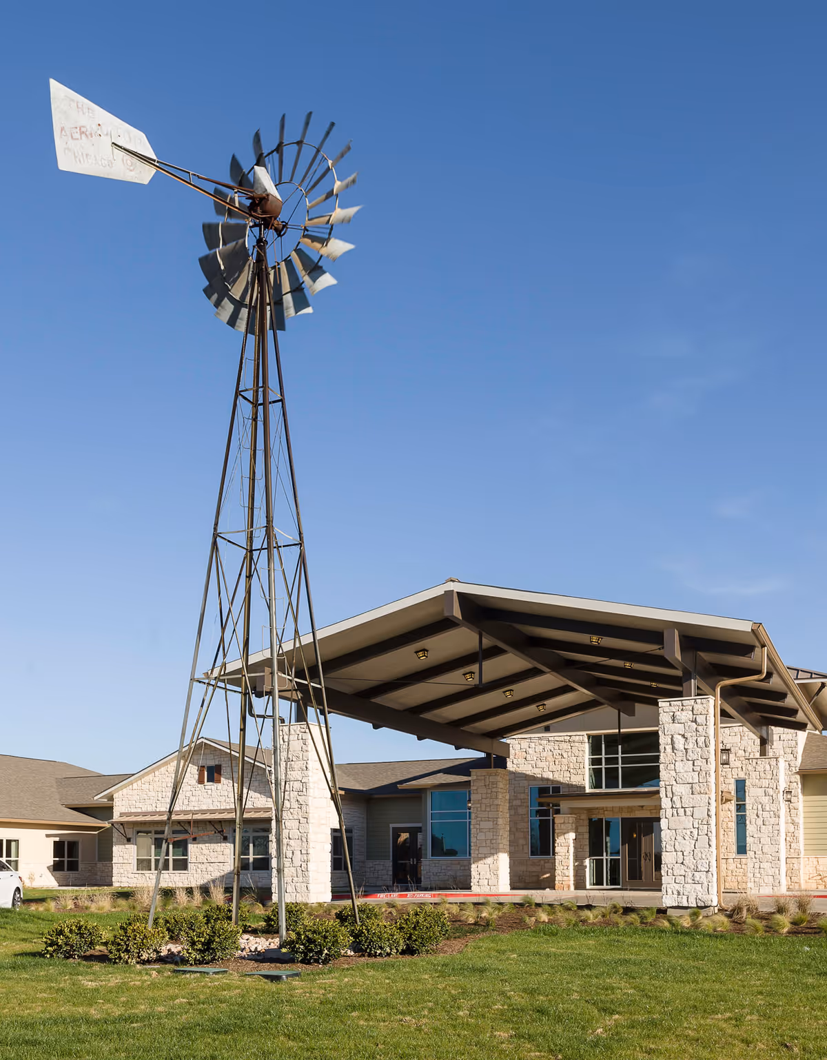 Exterior view of a senior living facility with a large metal windmill in the foreground, a stone and beige building with a covered entrance, green lawn, and clear blue sky.
