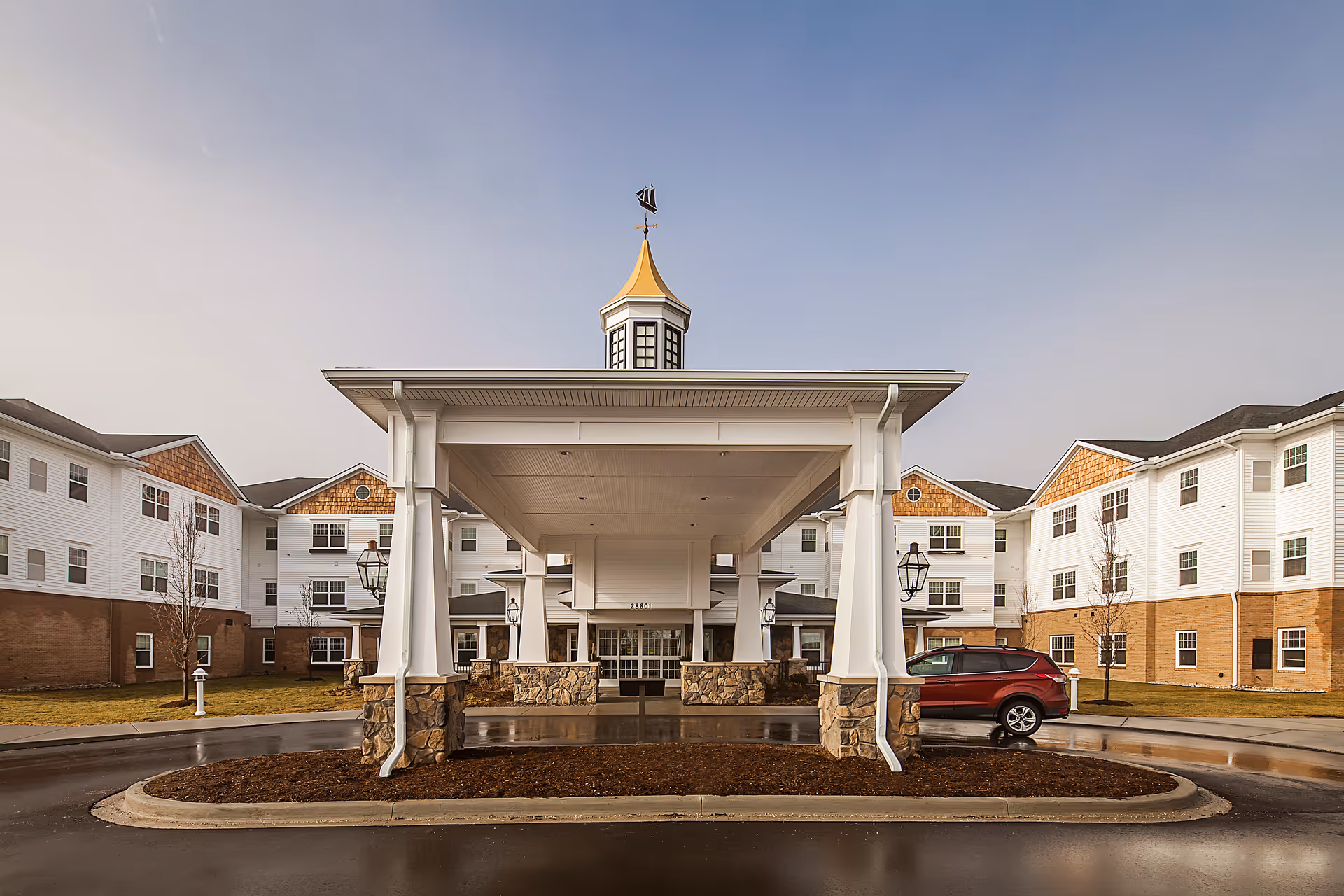 Front exterior view of American House Lakeshore senior living facility with a covered entrance featuring white columns and stone bases, a small cupola with a weather vane on top, and a red SUV parked nearby. The building has white siding with brown brick on the lower portion and multiple windows.