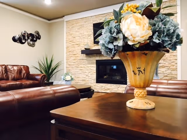 A cozy living room with brown leather sofas, a wooden coffee table with a decorative flower vase, a stone accent wall with a built-in fireplace, and a green potted plant in the corner.