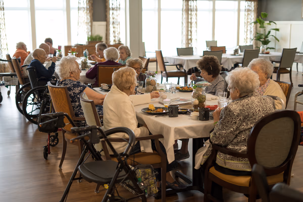 A group of elderly people sitting around a table in a dining room, eating and socializing. The room is well-lit with large windows and decorated with plants and curtains. Some individuals use wheelchairs and walkers.