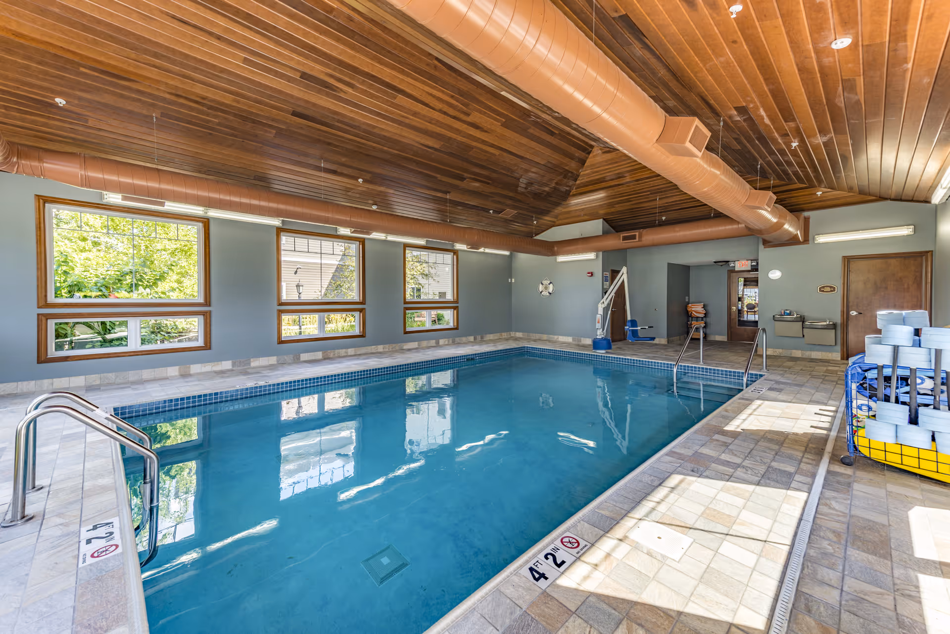 Indoor swimming pool at a senior living facility with a tiled deck, wooden ceiling, large windows, and a pool lift.