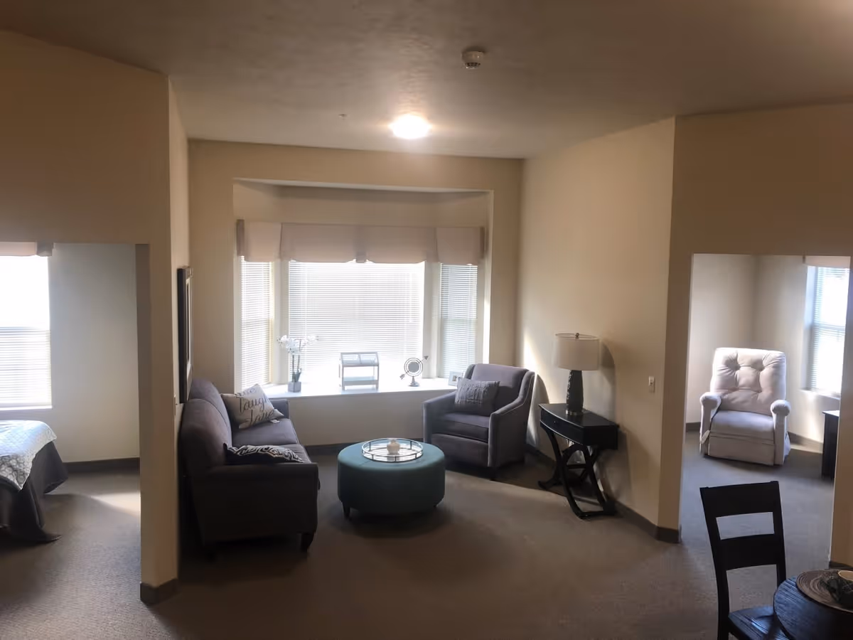 A cozy living area in an assisted living facility with a gray sofa, an armchair, a round ottoman, and a side table with a lamp. There is a large window with blinds and a small decorative fan on the windowsill. Adjacent rooms are visible, one with a bed and another with a cushioned chair near a window.