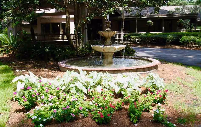 A circular stone water fountain surrounded by a garden bed with green and white foliage and colorful flowers, set in front of a building with a covered porch and trees providing shade.