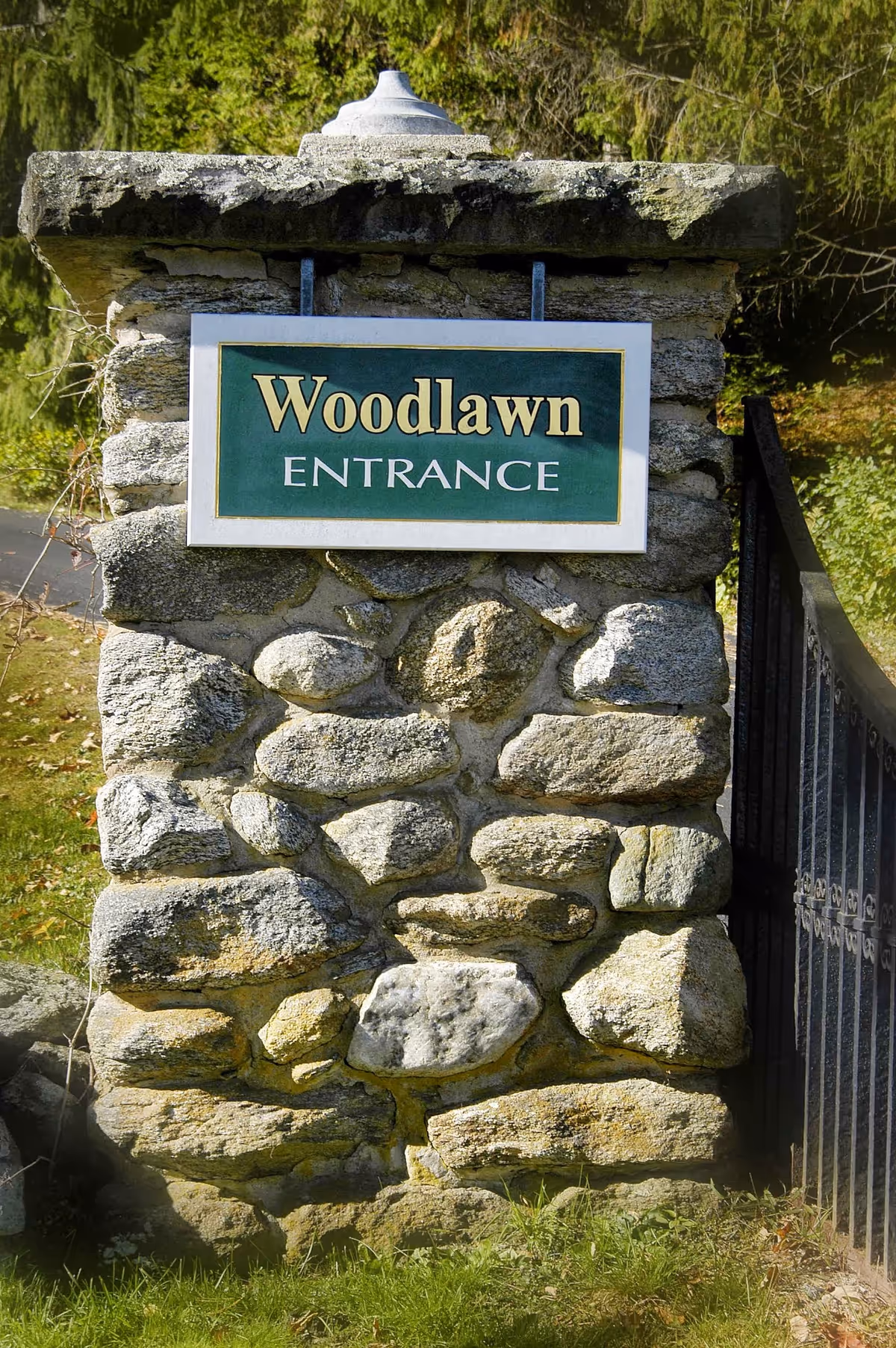 Stone entrance pillar with a green sign reading 'Woodlawn ENTRANCE' next to a metal gate and trees in the background.