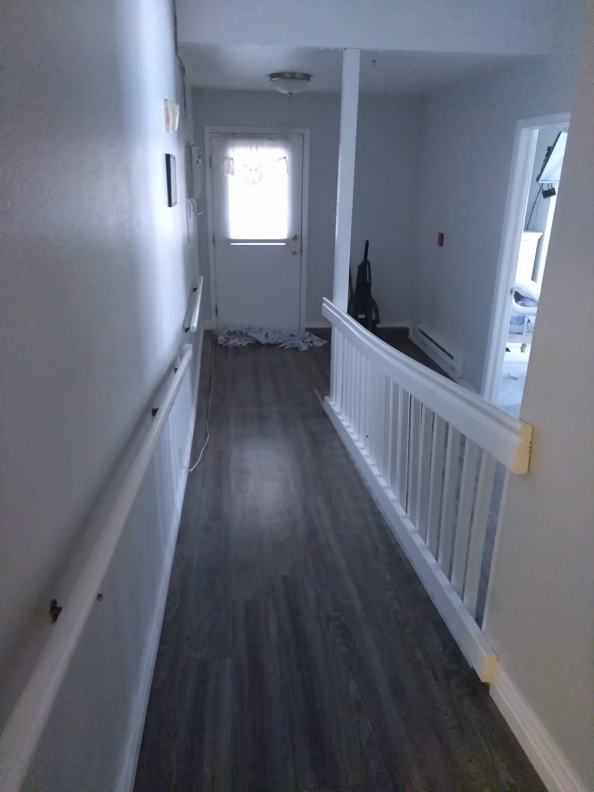 A narrow hallway with dark wood flooring and white walls. There are handrails on the left wall and a white railing on the right side. At the end of the hallway is a door with a window covered by a curtain, and a vacuum cleaner is placed near the corner. A room with a white dresser and a lamp is visible through an open door on the right.