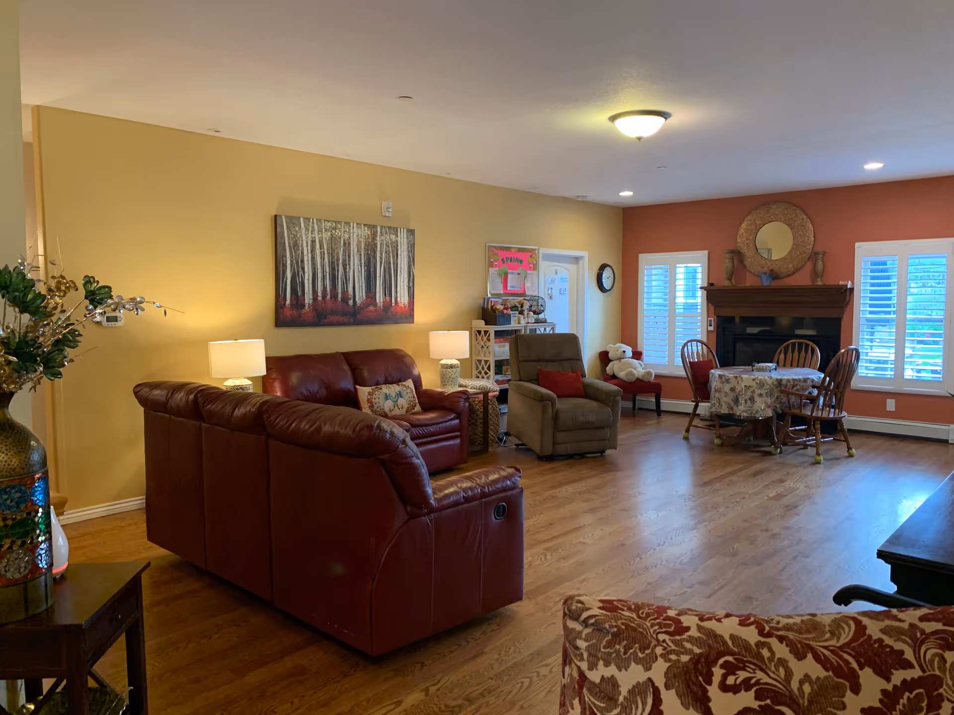 A cozy living room with wooden flooring, a red leather sectional sofa, a beige recliner chair, a round dining table with floral tablecloth and wooden chairs, a fireplace with a round mirror above it, two windows with white shutters, and a large painting of birch trees on a yellow wall.