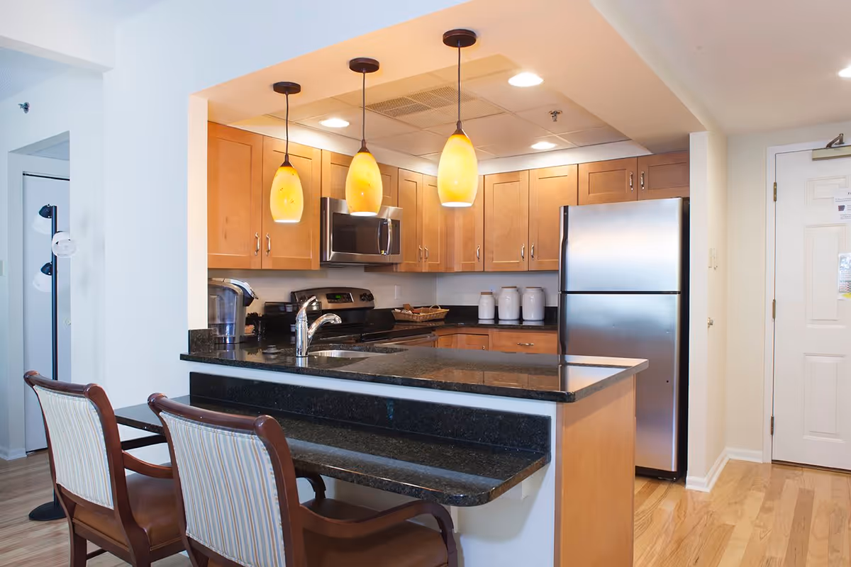 Modern kitchen area with wooden cabinets, black granite countertops, a stainless steel refrigerator, stove, and microwave. Three pendant lights with yellow shades hang above the counter, which has two wooden chairs with striped cushions. The floor is light wood, and there is a white door to the right.