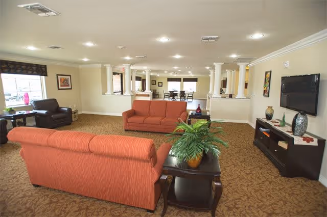 A spacious senior living facility common area with two orange sofas, a dark brown armchair, a side table with a potted plant, a TV mounted on the wall, and a cabinet below it. The room has beige walls, carpeted floor with a floral pattern, and several columns separating the seating area from a dining area in the background.