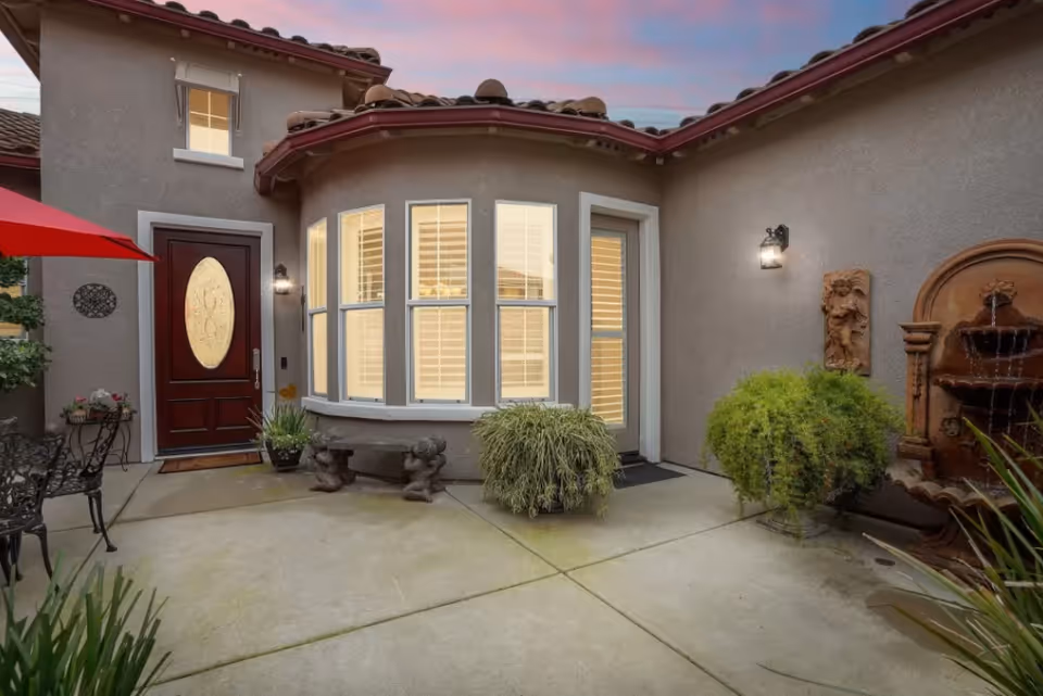 Outdoor courtyard area of a residential facility with a red front door, large bay windows with white shutters, potted plants, a stone bench, and a decorative water fountain on the right wall. The sky is visible with a pinkish hue at dusk.