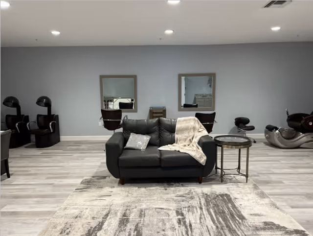 A small lounge with a black leather loveseat on a patterned rug, a side table, and salon chairs and hair dryers along a gray back wall.