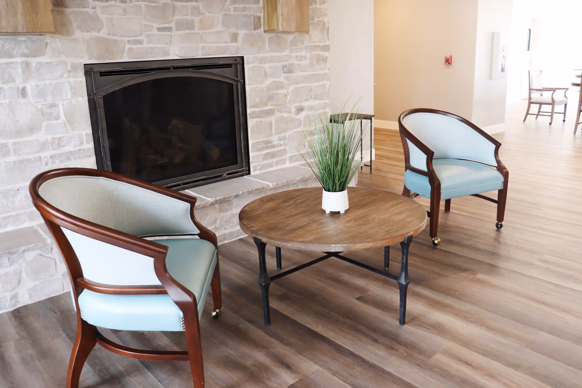 A cozy seating area with two light blue upholstered chairs with wooden frames positioned around a round wooden coffee table with a small potted plant on it, set in front of a stone fireplace in a room with wood flooring.