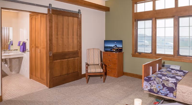 A senior living facility room with a hospital-style bed covered with a floral blanket, a wooden dresser with a TV on top, a striped armchair, large windows with wooden frames, and a sliding wooden door leading to a bathroom with a sink and mirror.