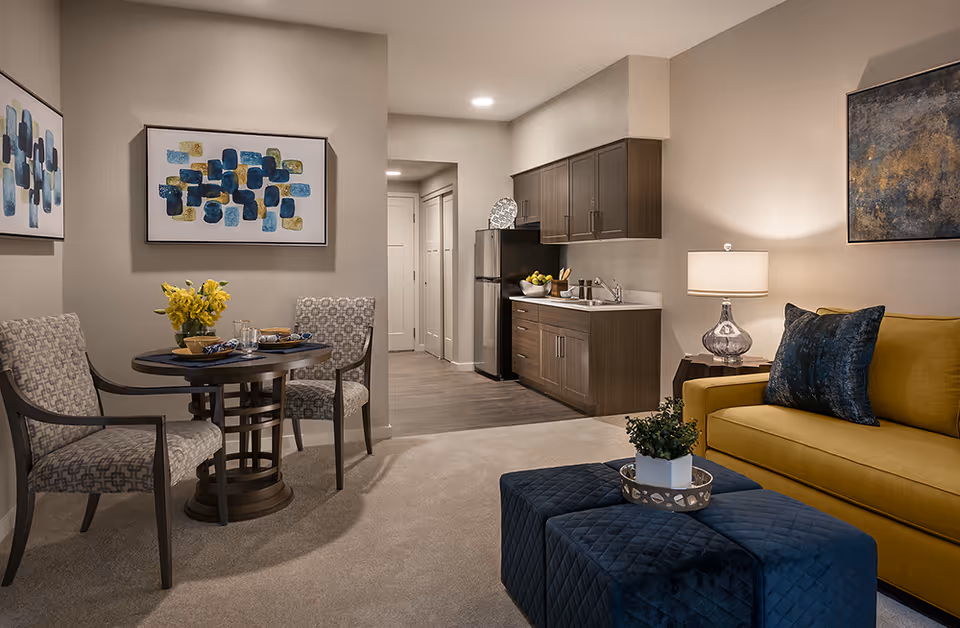 A cozy living area in a senior living facility featuring a yellow sofa with a blue pillow, a blue quilted ottoman with a small plant on a tray, a round dining table set for two with patterned chairs, and a kitchenette with wooden cabinets, a sink, and a refrigerator. The walls are decorated with abstract artwork, and the space is softly lit by a table lamp and recessed ceiling lights.