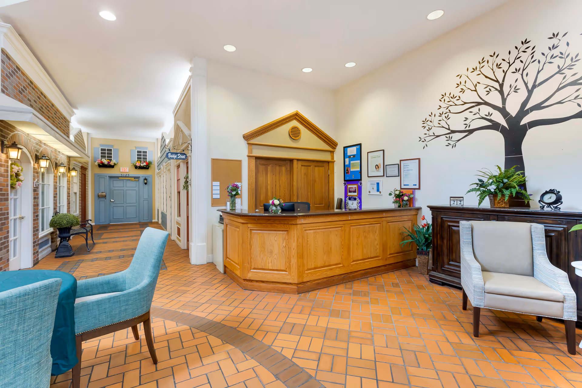 Reception area of a senior living facility with a wooden front desk, a large wall decal of a tree, framed certificates on the wall, and comfortable seating including a beige armchair and a teal chair around a table. The floor is tiled with a brick pattern, and the hallway features decorative storefront-style facades with windows and flower boxes.