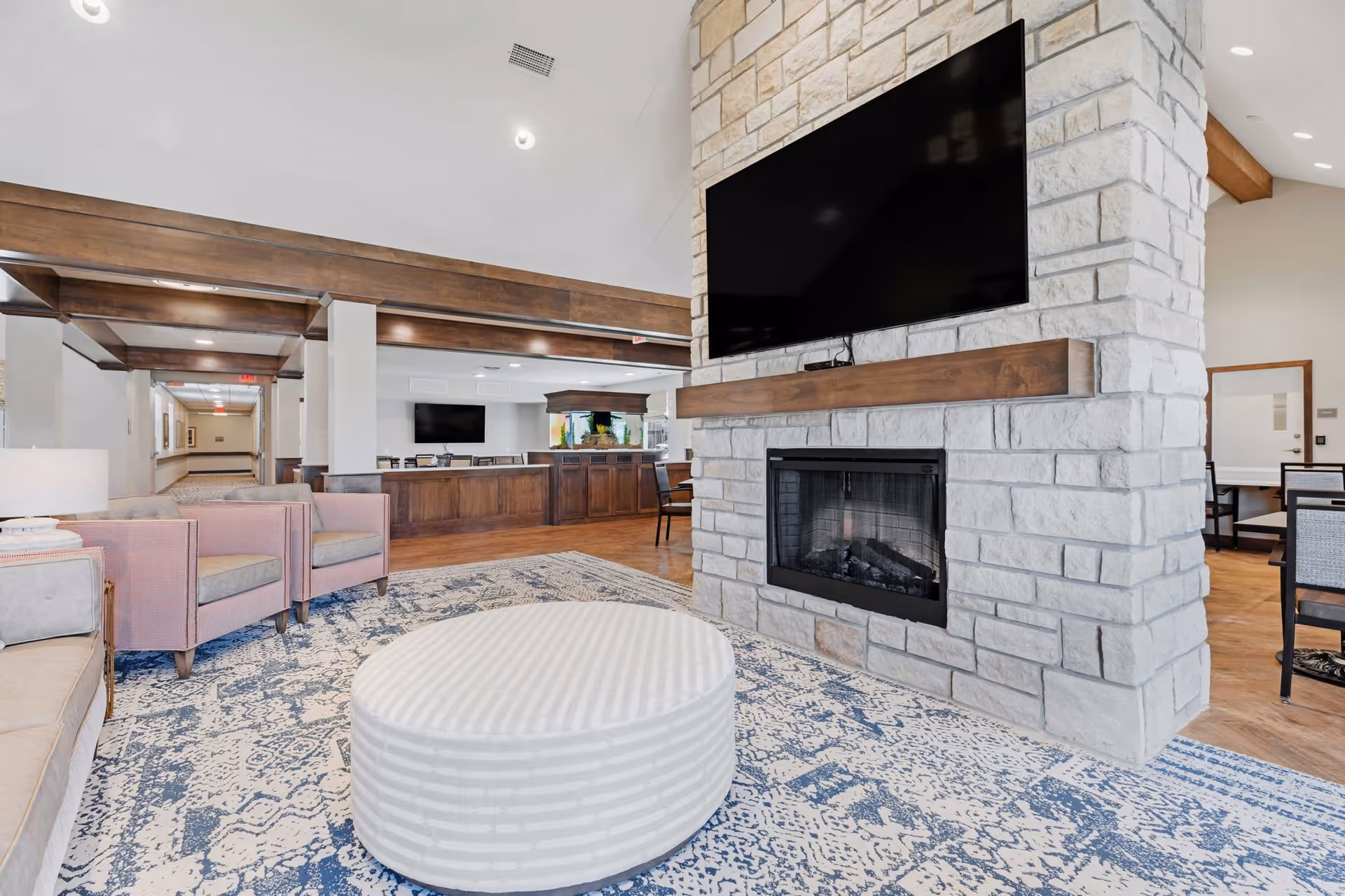 A spacious senior living common area featuring a large stone fireplace with a mounted flat-screen TV above it. The room has a patterned blue and white rug, a round striped ottoman, pink armchairs, and beige sofas. In the background, there is a wooden counter with an aquarium and additional seating, with wood beam accents on the ceiling.