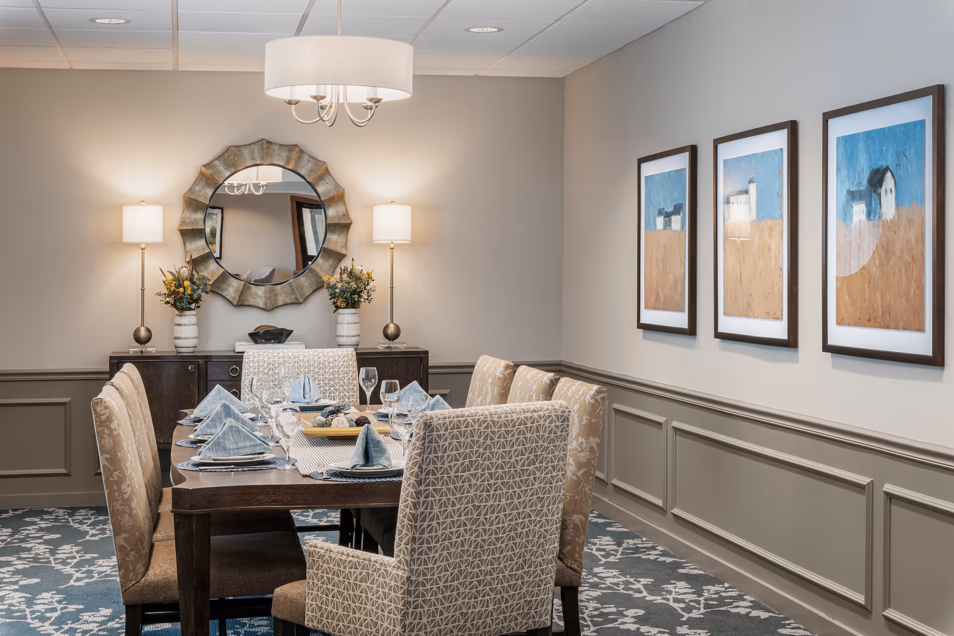 A neatly set dining table with upholstered chairs in a dining room featuring a sideboard, round mirror, lamps, and framed artwork on the wall.
