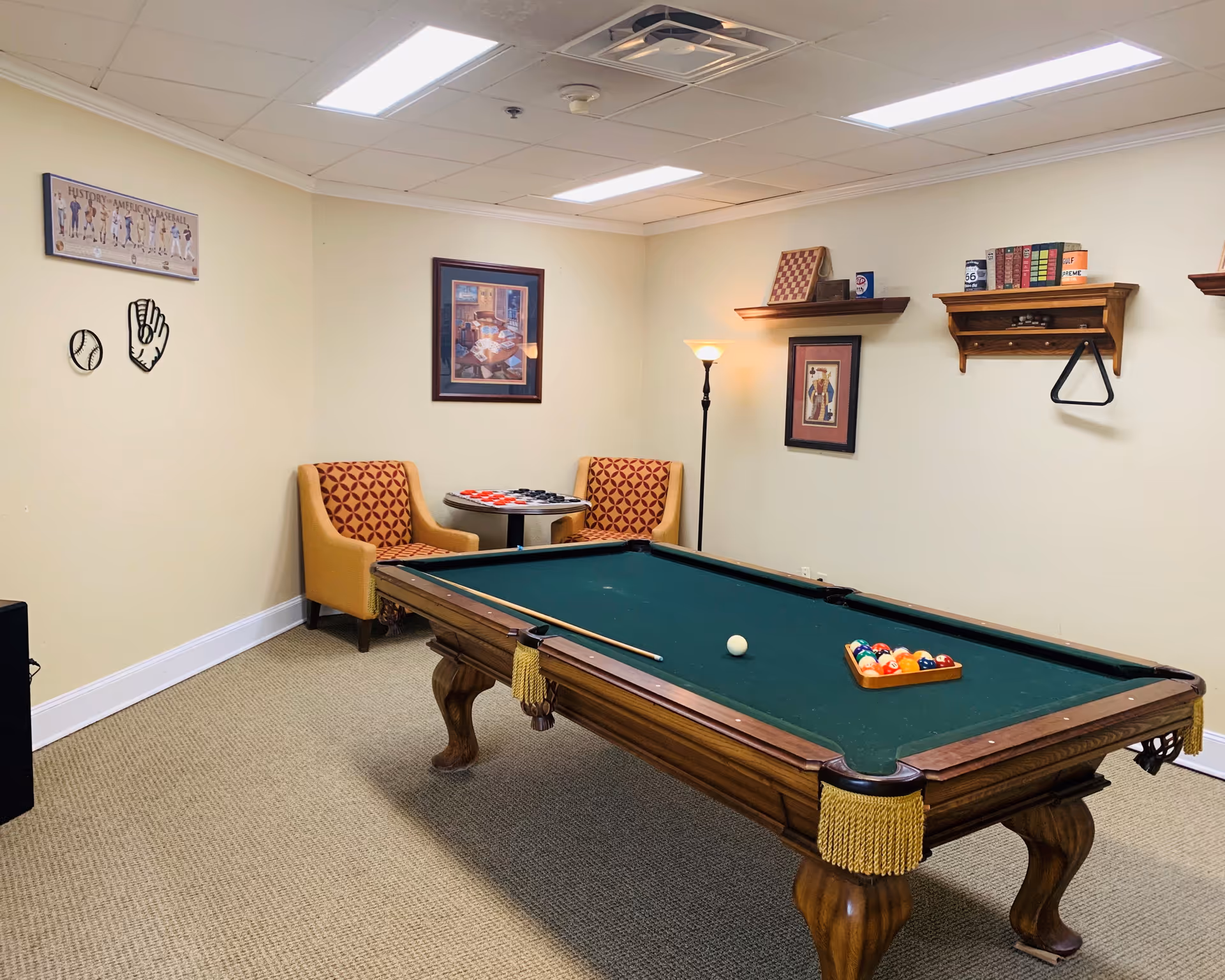 A recreational room with a pool table in the center, a cue stick and balls arranged on the table. Two patterned armchairs are placed near a small round table with a checkers board. The walls have framed pictures, shelves with books and decorative items, and a floor lamp providing light.
