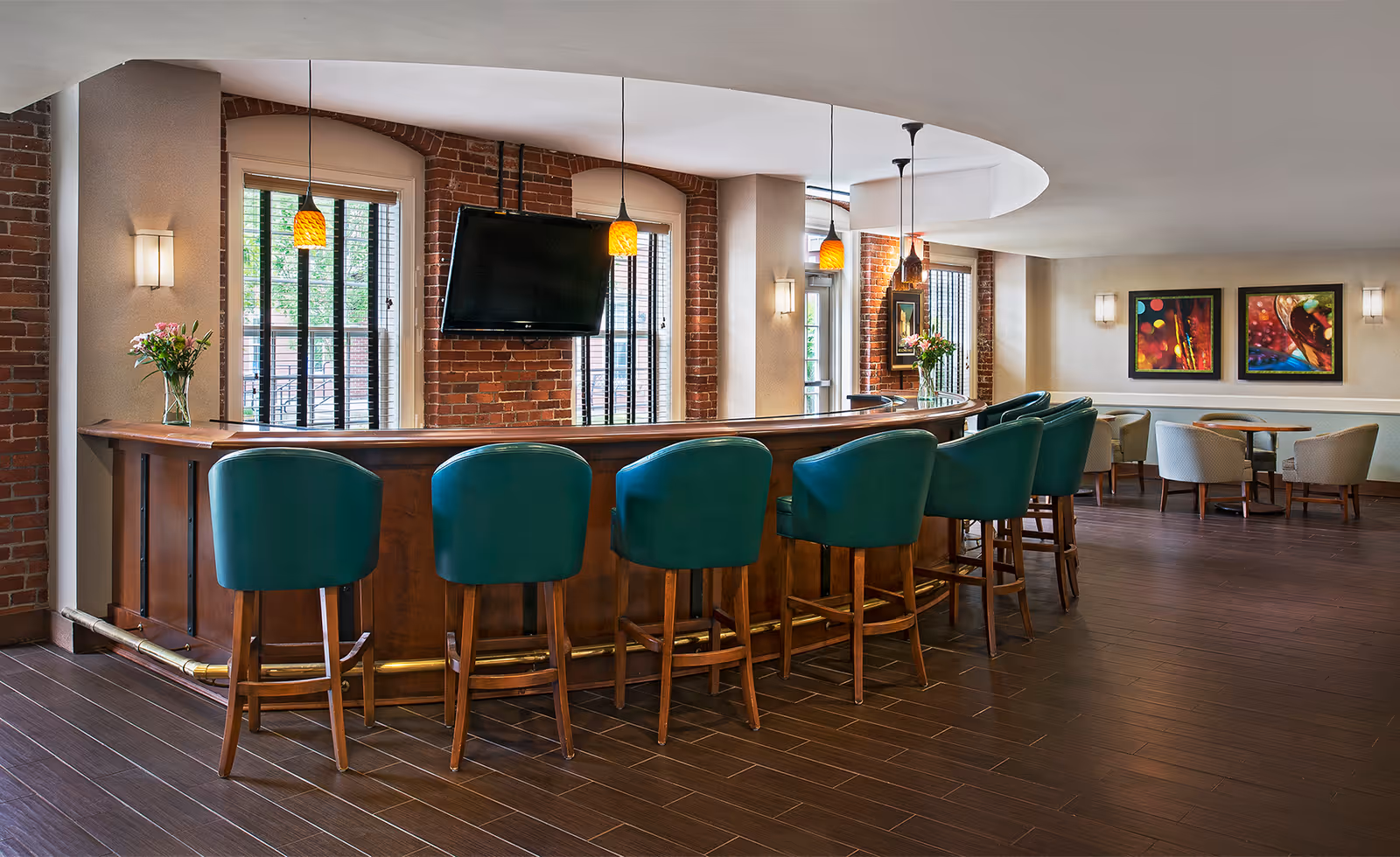 Interior view of a senior living facility lounge area featuring a curved wooden bar with six teal bar stools. The bar is decorated with two vases of flowers and illuminated by hanging pendant lights. Behind the bar are three large windows with black bars and a mounted flat-screen TV. In the background, there are several tables with chairs and colorful artwork on the walls.