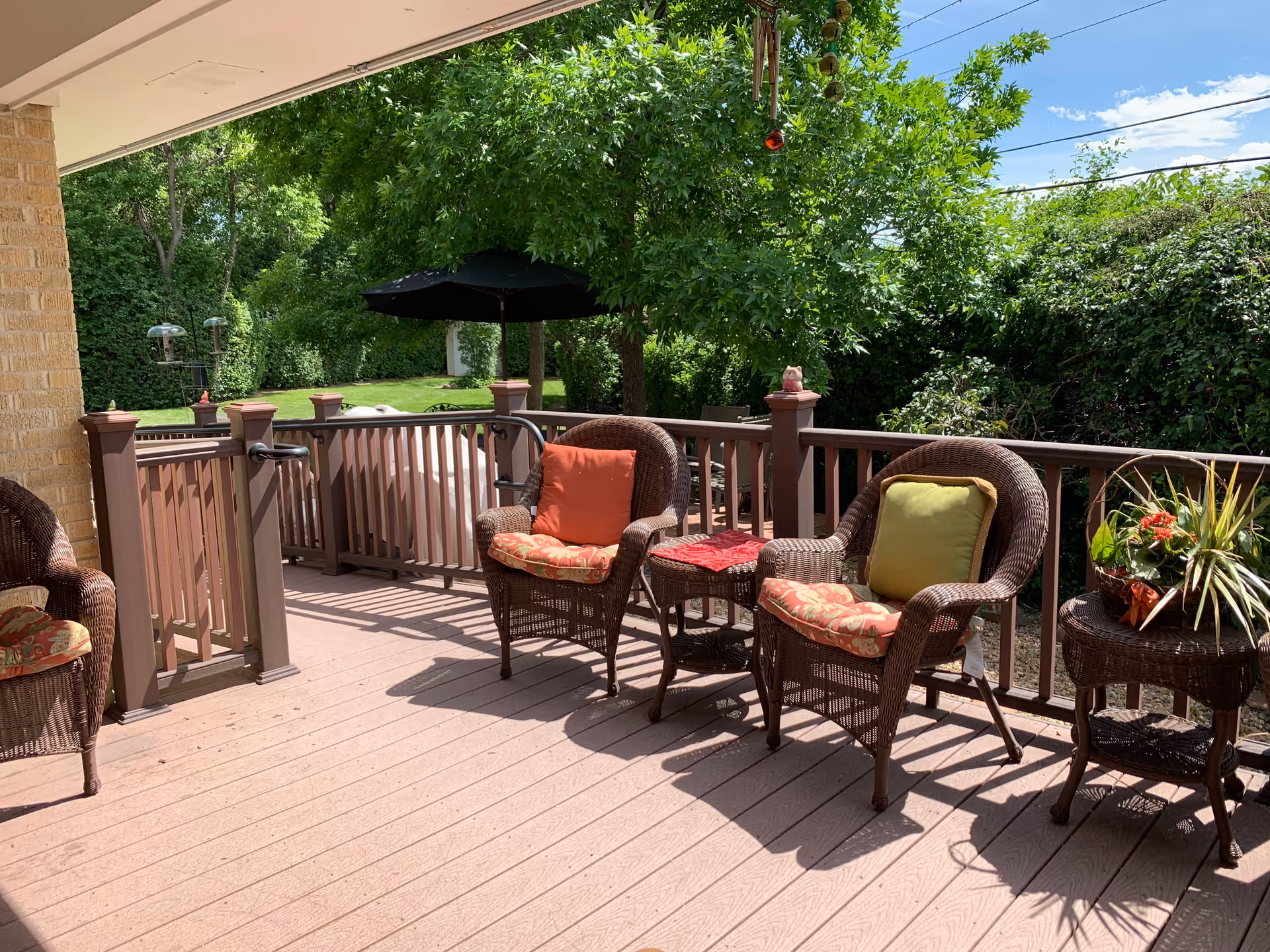 A shaded outdoor patio area with brown wicker chairs featuring colorful cushions, a small wicker table with a red cloth, and another wicker table with a potted plant. The patio overlooks a green garden with trees and bushes under a clear blue sky.