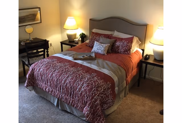 A cozy bedroom with a bed dressed in a red and beige comforter with white floral patterns, multiple pillows, and a tray on top. There are two bedside tables, each with a lamp, and a small plant on one table. A chair and desk with a mirror and framed artwork are visible in the corner.