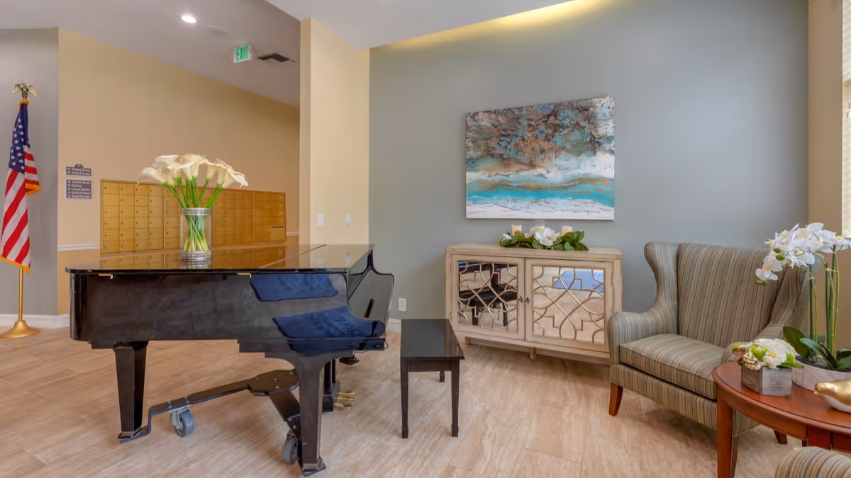 A cozy interior lounge area featuring a black grand piano with a vase of white flowers on top, a striped armchair next to a wooden side table with a white orchid plant, and a mirrored cabinet with decorative greenery and candles. An abstract painting with blue and brown tones hangs on the gray wall behind the cabinet. An American flag stands in the corner near a wall with mailboxes.