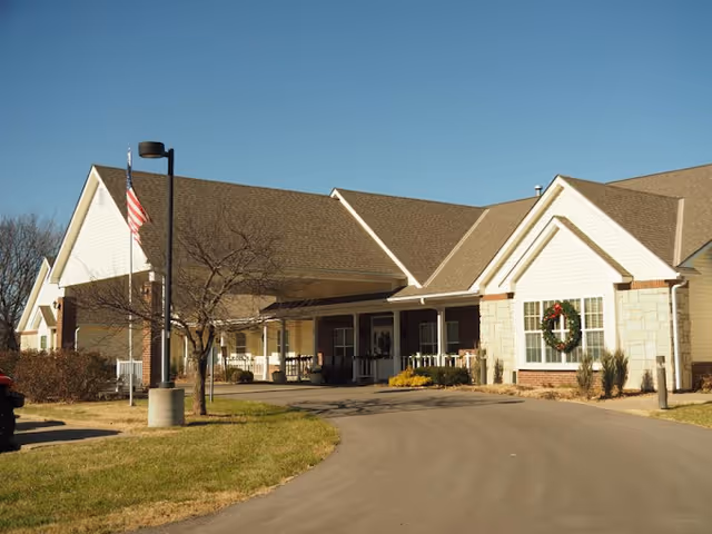 Exterior view of a single-story senior living facility building with a covered entrance, a wreath on the front window, an American flag on a pole, and a clear blue sky.