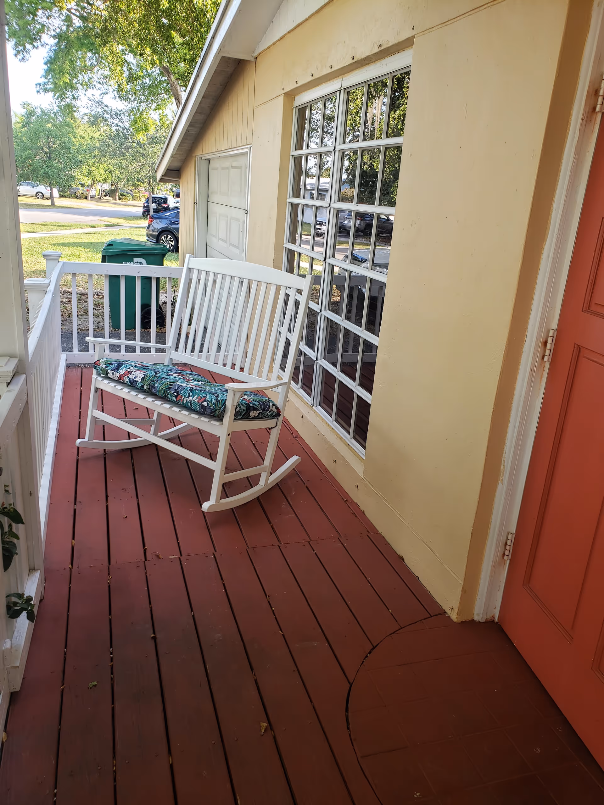 A small porch area with reddish wooden flooring, a white rocking bench with a floral cushion, a large window with multiple panes, a beige exterior wall, and a red door. In the background, there are green trash bins and some parked cars visible on the street.