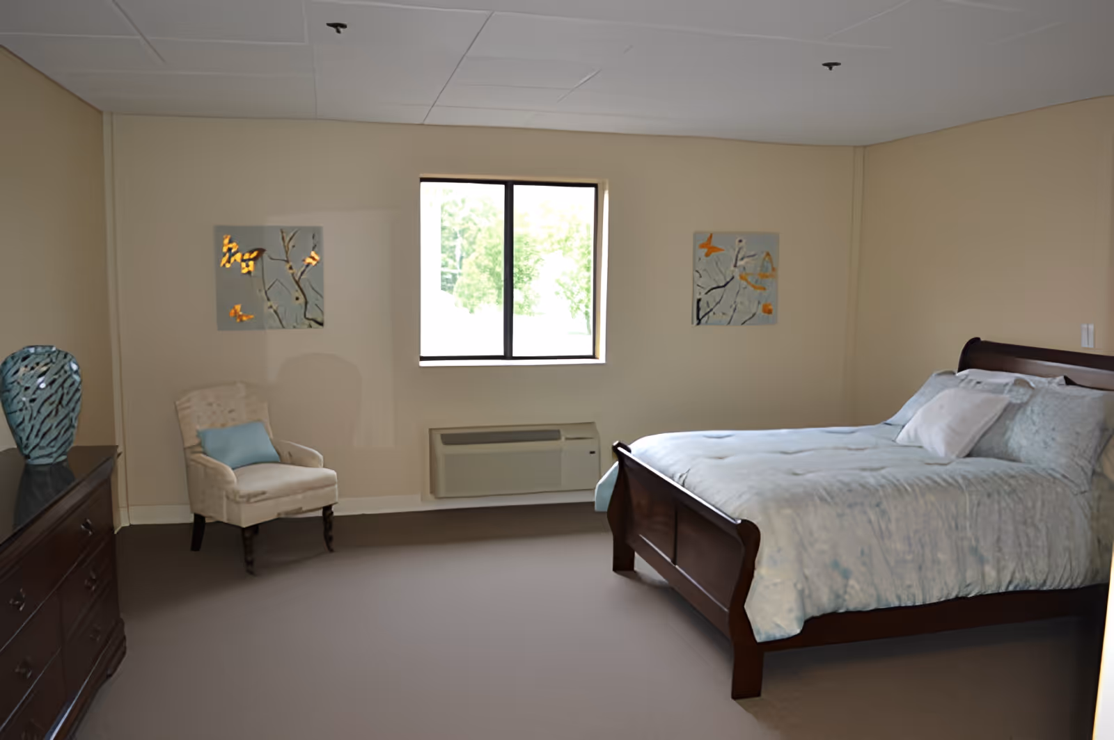 A senior living bedroom with a wooden bed frame and light blue bedding, a beige armchair with a blue cushion, a wooden dresser with a decorative blue vase, two paintings on the wall, and a window letting in natural light.