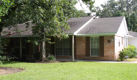 Single-story brick residential building with a sloped roof, surrounded by green grass and trees. The building has several windows with brown shutters and a small covered porch area.