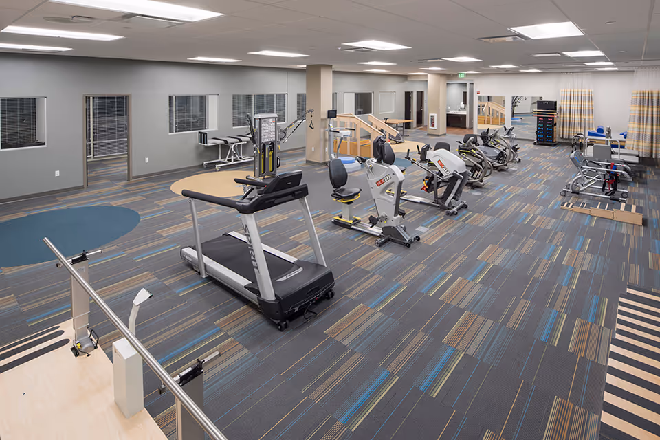 A spacious fitness room in a senior living facility featuring various exercise equipment including a treadmill, stationary bikes, and strength training machines. The room has patterned carpet flooring, neutral-colored walls, large windows, and bright overhead lighting. There are also parallel bars and a small staircase for physical therapy exercises.