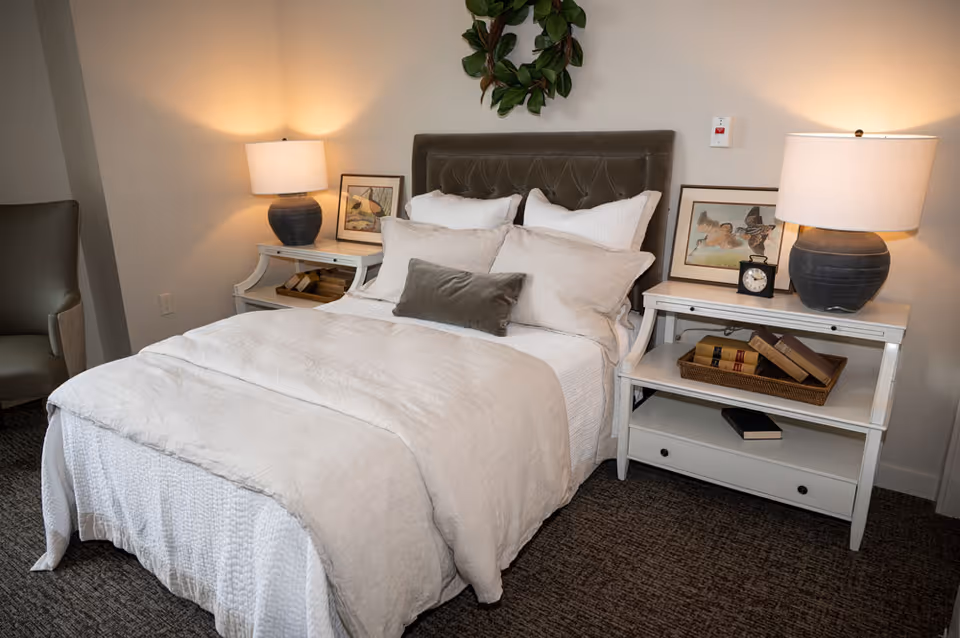 A neatly made bed with white and beige bedding, flanked by two white nightstands each with a lamp, framed artwork, and books. A green armchair is partially visible to the left, and a decorative wreath hangs above the bed's headboard.