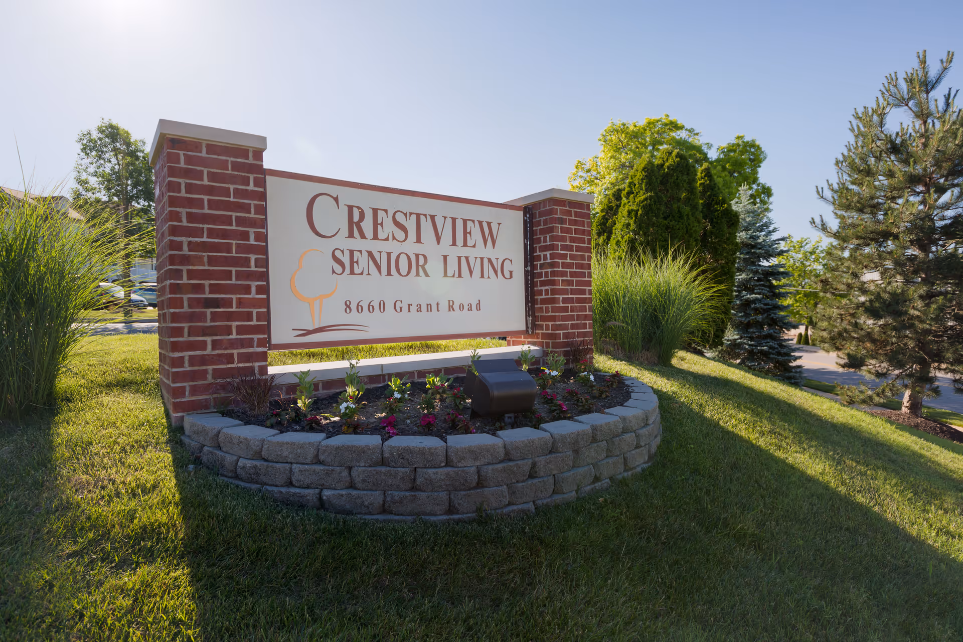 Outdoor view of a brick and stone sign for Crestview Senior Living located at 8660 Grant Road, surrounded by grass, plants, and trees under a clear sky.