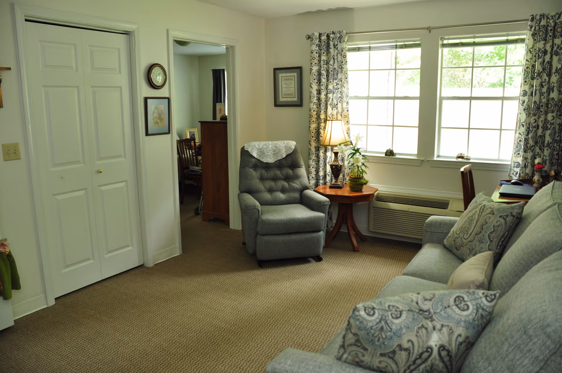 A cozy living room area with a gray upholstered recliner chair next to a wooden side table holding a lamp and a potted plant. There is a large window with patterned curtains allowing natural light to fill the room. A gray sofa with patterned cushions is partially visible on the right. The room has beige carpet flooring and white walls with framed pictures and a clock. A doorway leads to another room with wooden furniture and a chair.
