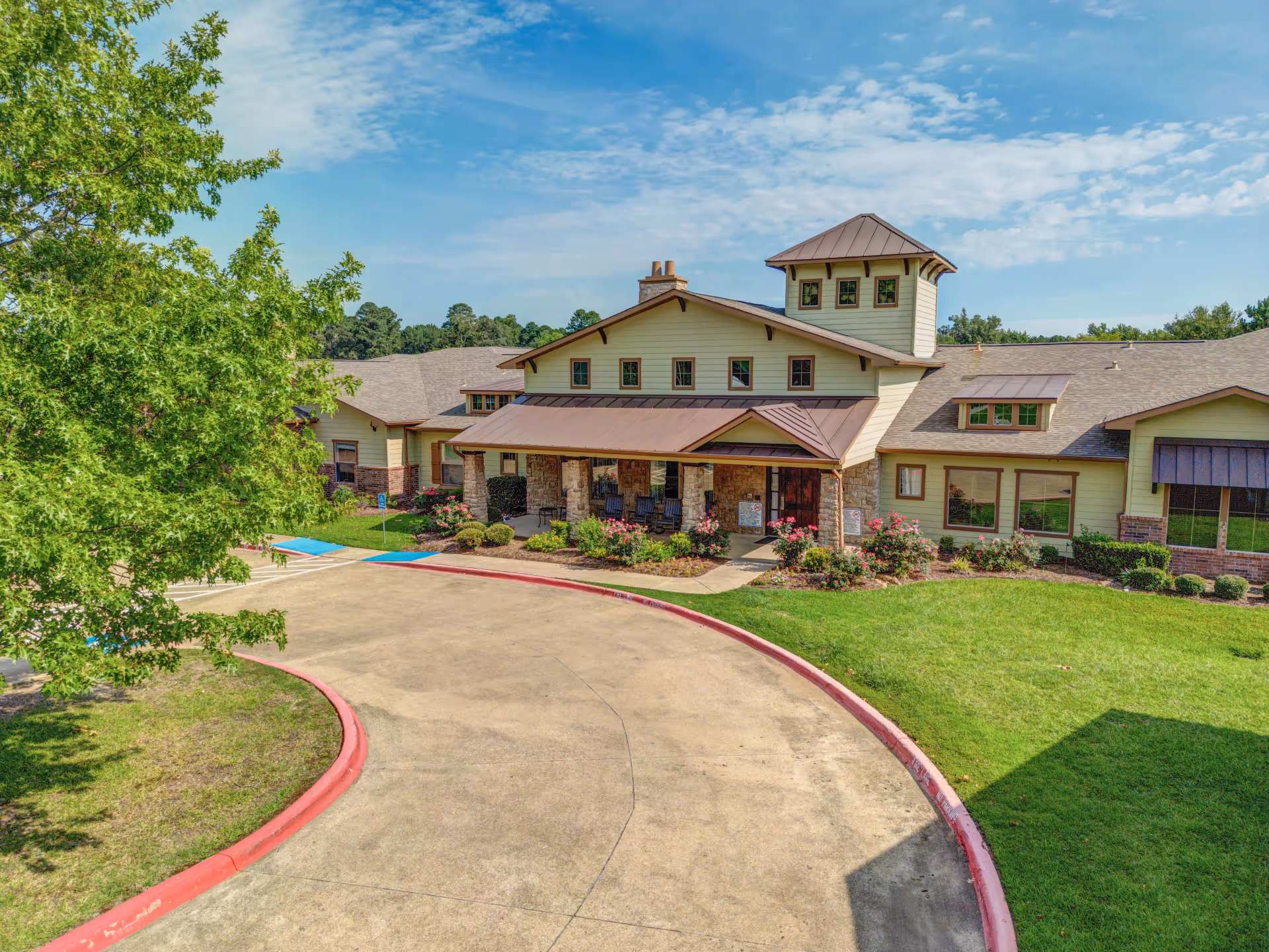 Exterior view of Hawkins Creek Assisted Living and Memory Care building with a covered entrance, stone pillars, and surrounding landscaping including green grass, bushes, and a tree. The sky is partly cloudy and blue.