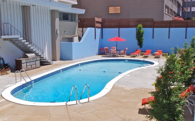 Outdoor swimming pool area with a kidney-shaped pool surrounded by a concrete deck. There are red lounge chairs and a table with a red umbrella near the pool. A staircase and a barbecue grill are visible on the left side, and some greenery is on the right side. The area is enclosed by a white wall and a wooden fence.