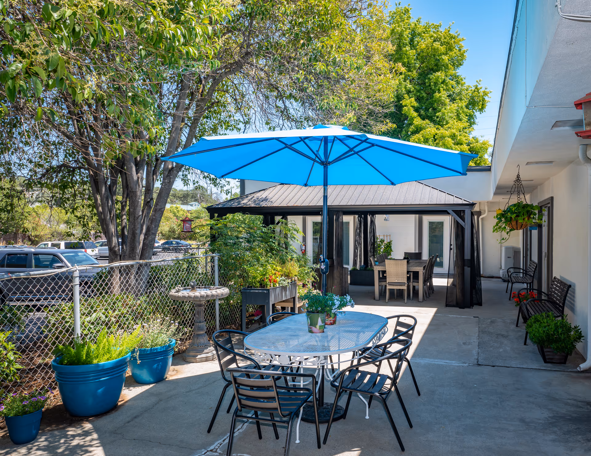 Outdoor patio area with a blue umbrella shading a glass table surrounded by black chairs. There are potted plants, a birdbath, and a covered seating area with a dining table and chairs. Trees and parked cars are visible in the background.