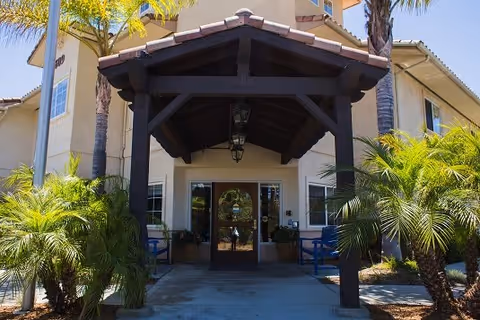 Entrance of a senior living facility named Regency Fallbrook with a covered walkway supported by dark wooden beams, surrounded by palm trees and greenery under a clear blue sky.