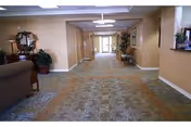 Spacious carpeted hallway in a senior living facility with seating areas, potted plants, and a reception desk leading to glass exit doors.