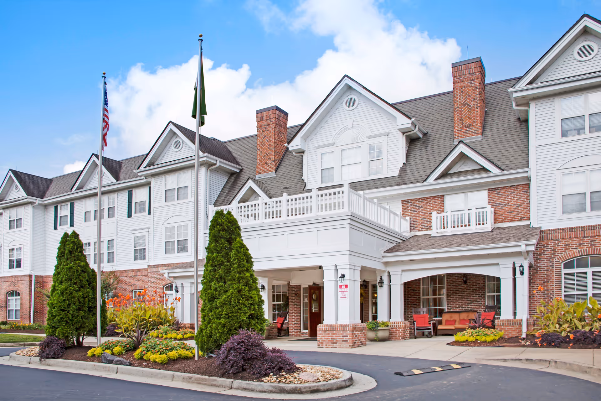 Front exterior view of PruittPlace - Buckhead, a multi-story senior living facility with white siding and red brick accents. The entrance features a covered drop-off area with columns, outdoor seating with red cushions, and landscaped greenery including tall shrubs and flower beds. Two flagpoles with the American flag and another flag stand near the entrance under a partly cloudy sky.