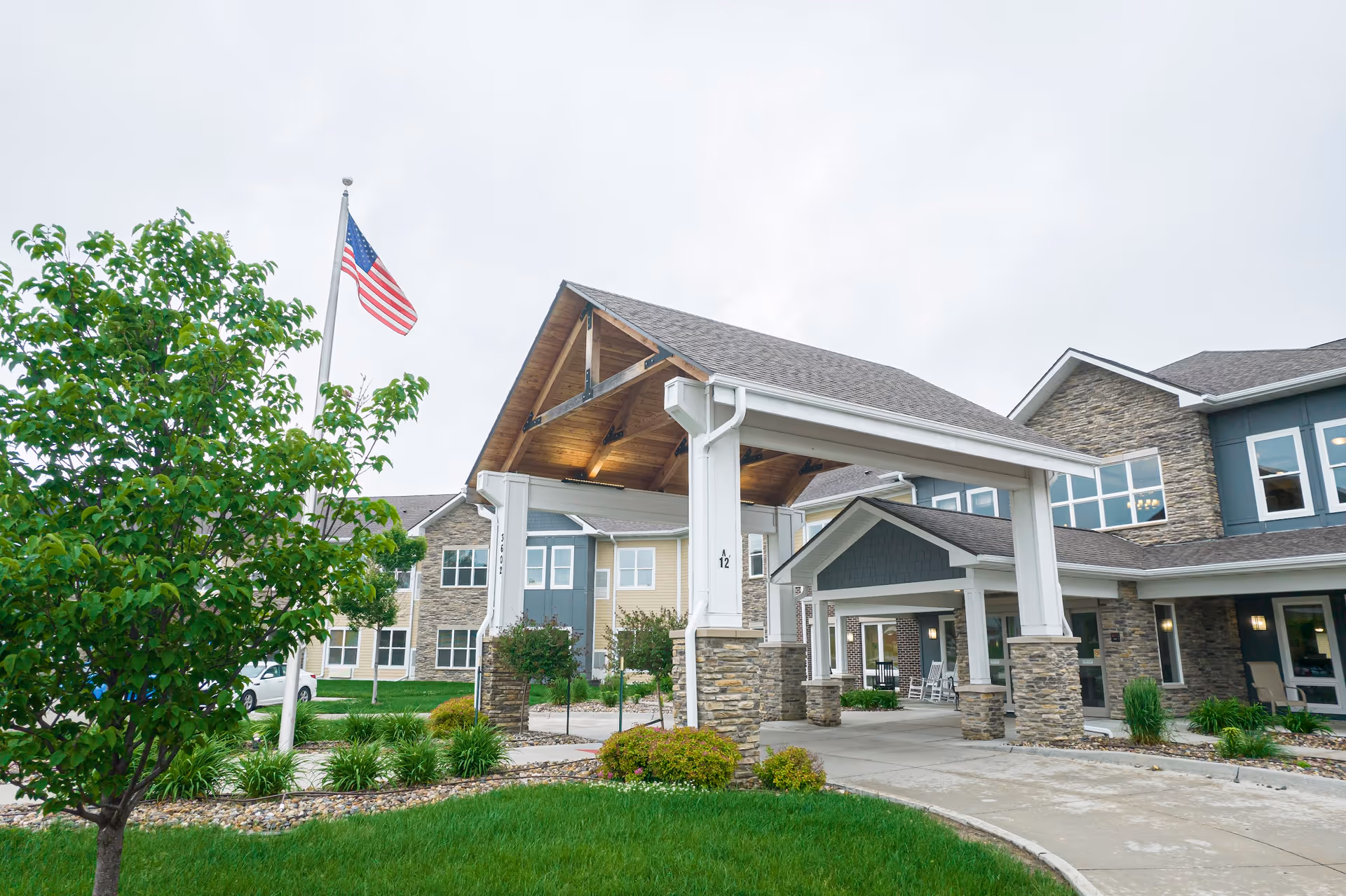 Covered entrance and driveway of a senior living building with an American flag, stone columns, and landscaped lawn.