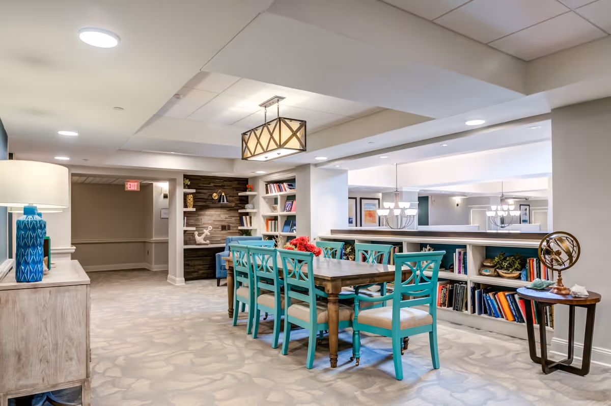 A bright and spacious common area in a senior living facility featuring a long wooden table surrounded by turquoise chairs. The room has built-in bookshelves filled with books and decorative items, a blue lamp on a sideboard, and modern light fixtures on the ceiling. The carpet has a subtle pattern, and the walls are painted in neutral tones.