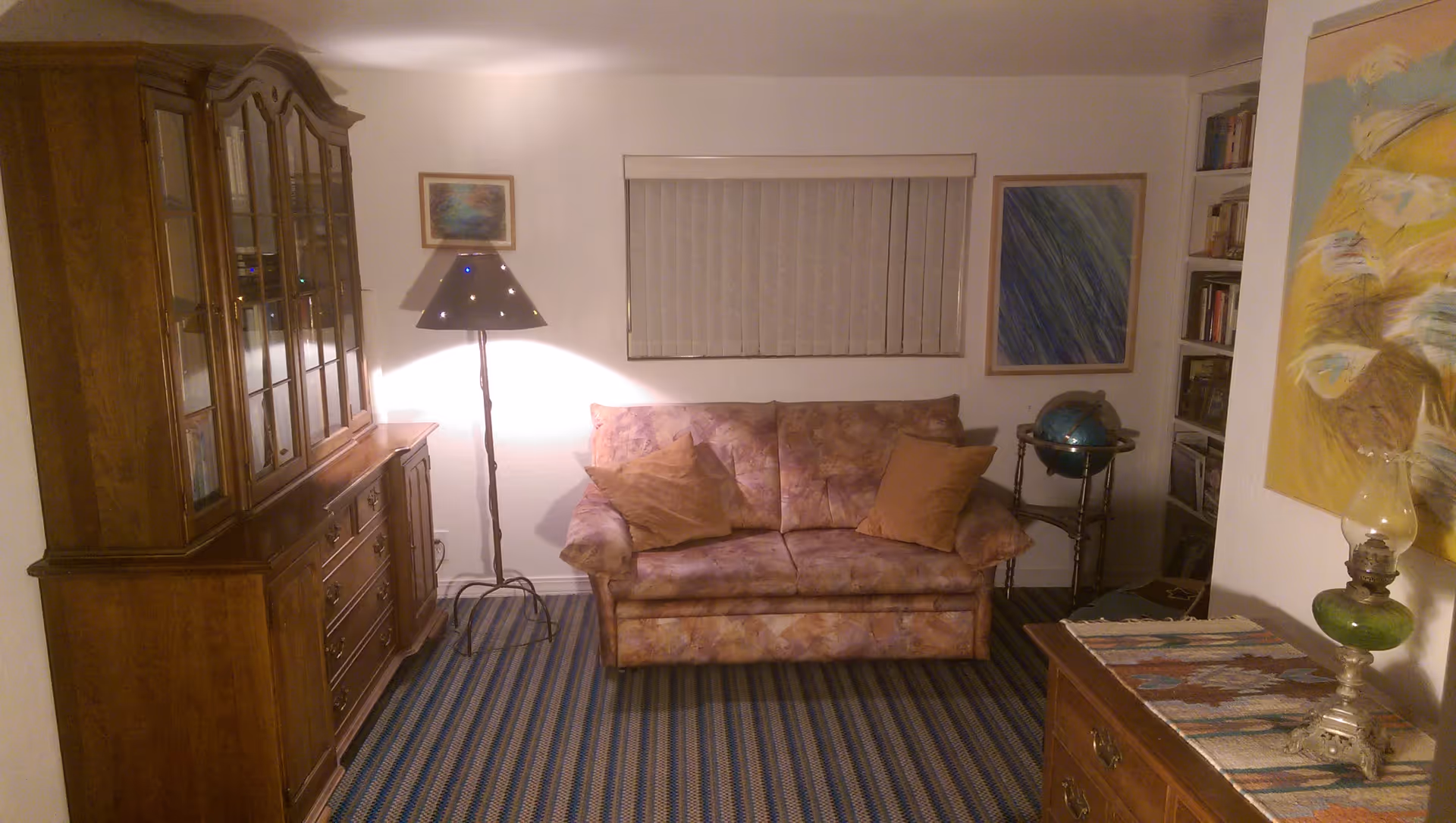 A cozy living room with a patterned sofa and two brown cushions, a tall floor lamp with a perforated shade, a wooden cabinet with glass doors on the left, a globe on a small stand, bookshelves, and framed artwork on the walls. The floor is covered with a striped carpet.