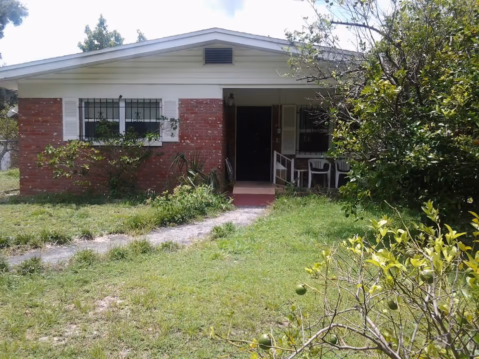 Front exterior view of a single-story brick house with white trim and a small covered porch. The porch has two white plastic chairs and a small table. There is a grassy yard with some bushes and trees in front of the house.