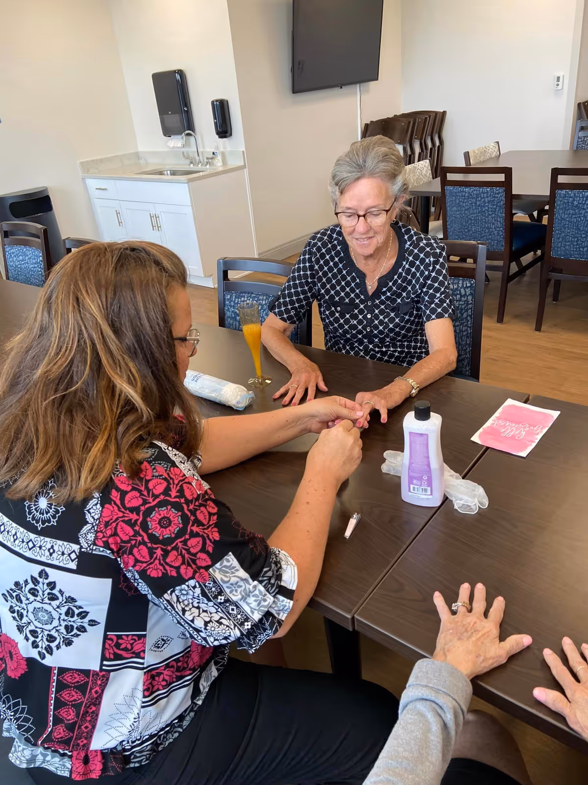 Two women sitting at a table in a common room, one giving the other a manicure. The room has wooden floors, a sink with cabinets, a wall-mounted TV, and several chairs and tables. A bottle of lotion and manicure tools are on the table.