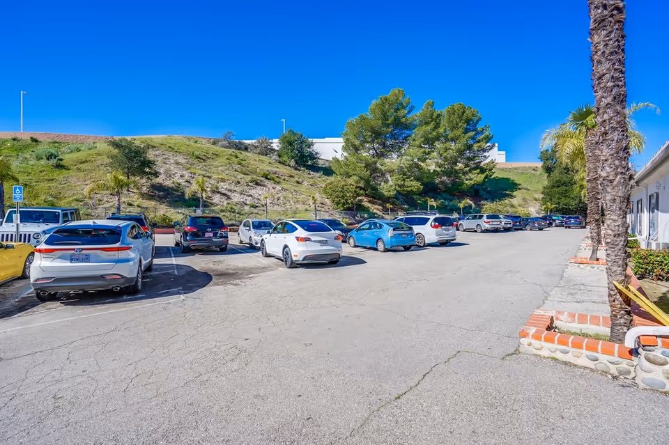 Parking lot with multiple parked cars next to a building, with a hillside covered in grass and trees in the background under a clear blue sky.